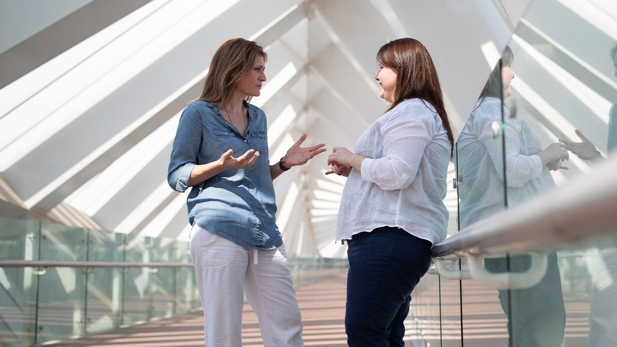 Two women engaged in a professional coaching conversation in a bright, modern architectural hallway.