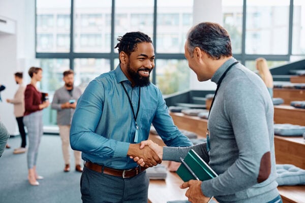 Two men are shaking hands in an casual networking environment while smiling at each other.