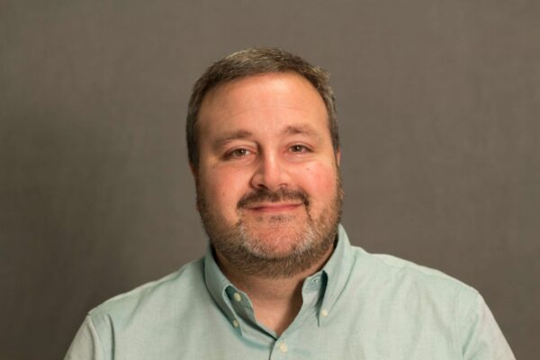 Mark Ruth headshot photo, smiling man wearing light green button up shirt in front of tan background