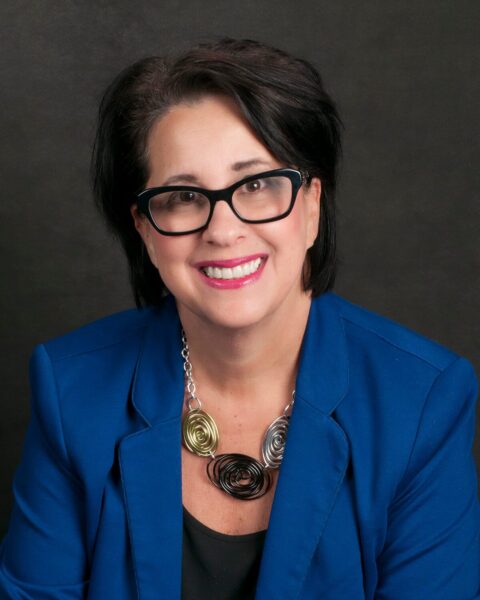 Barb Girson PCC ACC headshot photo, smiling woman wearing blue blazer black top and metal necklace in front of dark gray background