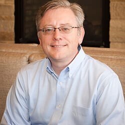 Matt Barney headshot photo, smiling man with gray hair wearing glasses and a light colored collared shirt in front of fireplace