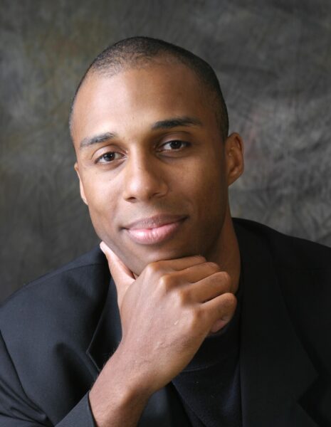 Brian Hillard headshot photo, smiling man wearing black suit in front of gray background