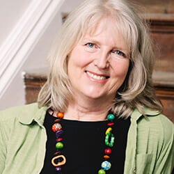 Jane Brottchie CPCC ACC PCC headshot photo, woman smiling with white hair wearing a green shirt over a black shirt and colorful necklace in front of a stairway