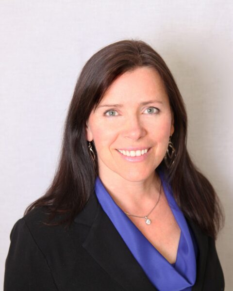 Catherine Carr headshot photo, smiling woman with dark hair wearing a black blazer and purple top in front of a white wall