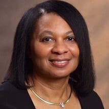 Dr. Ormella Cummings ACC headshot photo, smiling woman with dark hair wearing a gold necklace and dark top in front of a brown background