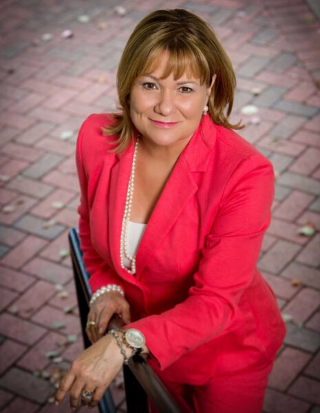 Darelyn DJ Mitsch MCC headshot photo, smiling woman wearing red suit leaning against a handrail outside