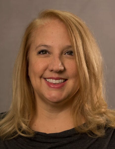 Amy Friskney headshot photo, smiling woman with blond hair wearing dark top in front of tan background