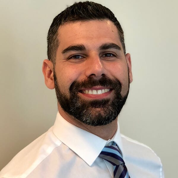 Joseph Grech ACC PCC headshot photo, smiling man with dark hair wearing white collared shirt and stripped tie in front of white wall