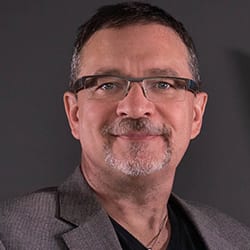 Greg Hadubiak MHSA FACHE PCC headshot photo, smiling man wearing glasses and a dark suit in front of a dark gray background