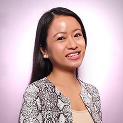 Joyce Talag ACC PCC headshot photo, smiling woman with dark hair standing in front of a pink background