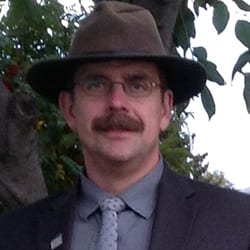 James Greengrass PCC headshot photo, smiling man with hat and glasses wearing a blazer collared shirt and tie outside