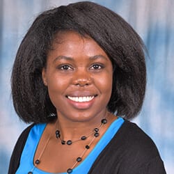 Kathleen Lihanda headshot photo, smiling woman with dark hair wearing a black beaded necklace black sweater and blue top in front of a light blue background