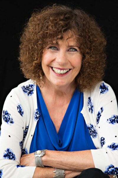 Kathy Harman MCC headshot photo, smiling woman wearing white and blue floral sweater over a blue top with arms crossed over her chest in front of black background