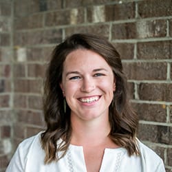 Laura Shrake headshot photo, smiling woman with brown hair and a white top standing in front of a brick wall