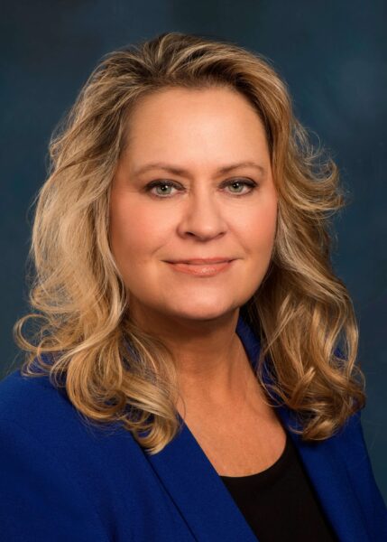 Dr Lynn Schmidt ACC headshot photo, smiling blond woman wearing blue blazer and black top in front of gray background