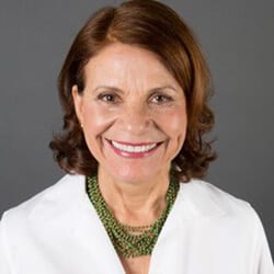 Merle McKinley MCC headshot photo, smiling woman with reddish brown hair wearing a green necklace and white top in front of a gray background