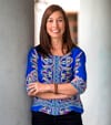Michele Oelking PCC headshot photo, smiling woman with long hair wearing a blue top arms crossed over her torso, standing against a white wall