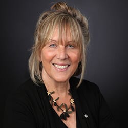 Cinnie Noble PCC headshot photo, smiling blond woman wearing dark top in front of dark gray background