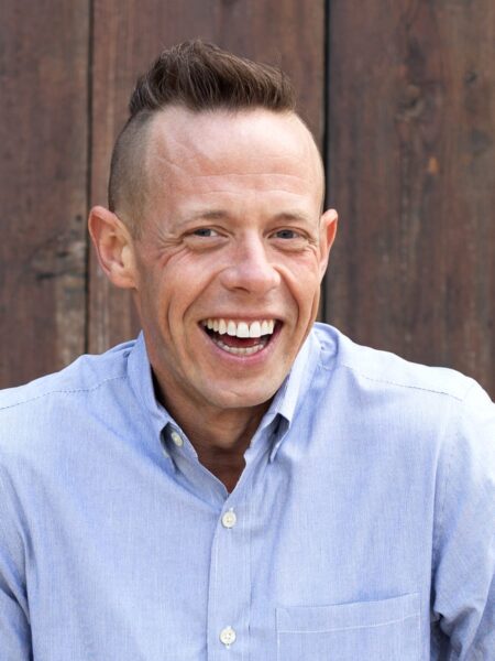 Patrick Burlingham headshot photo, smiling man wearing light blue button up shirt in front of wood wall