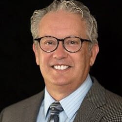 Paul White PCC headshot photo, smiling man with gray hair wearing glasses and dark brown suit in front of a black background