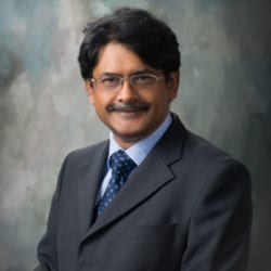 Prasad Deshpande MCC headshot photo, smiling man with dark hair wearing glasses and grey suit blue tie in front of neutral background