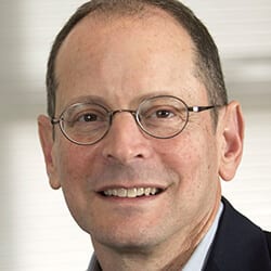 Jonathan Rauch headshot photo, smiling man wearing glasses and black blazer in front of neutral background