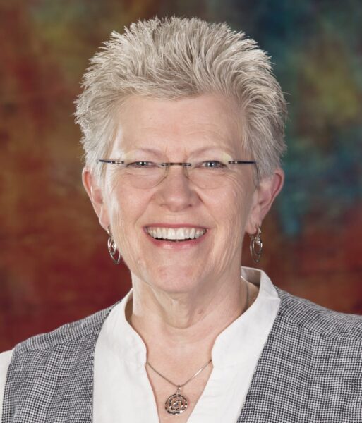 Sara Smith MCC headshot photo, smiling woman with gray hair wearing a patterned top in front of a multicolored background