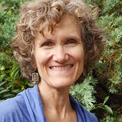 Dee Wagner headshot photo, woman smiling with curly brown hair and blue top in front of greenery