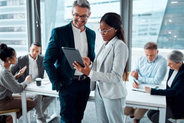 Two professionals reviewing a tablet during a business coaching session, with a group of colleagues collaborating in the background.
