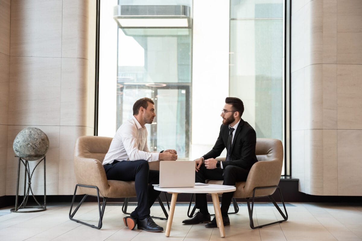 two men in discussion at a small side table with a laptop