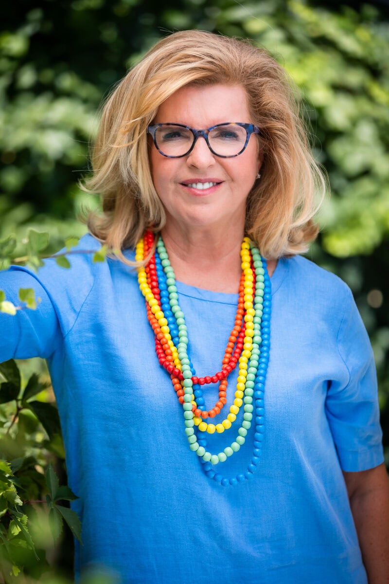 Judit Abri von Bartheld MA MCC ACTC headshot photo, smiling blond woman wearing blue top and multicolored necklaces outside