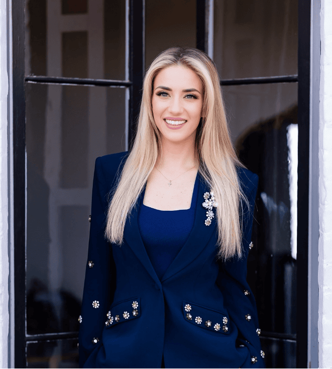 Casey Hughes headshot photo, smiling blond woman wearing navy blazer and dress in front of window