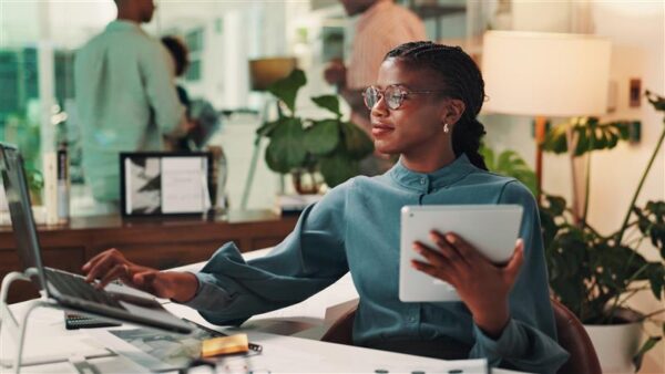 Professional Black woman in teal blouse working at desk with laptop and tablet in modern office with plants and colleagues.