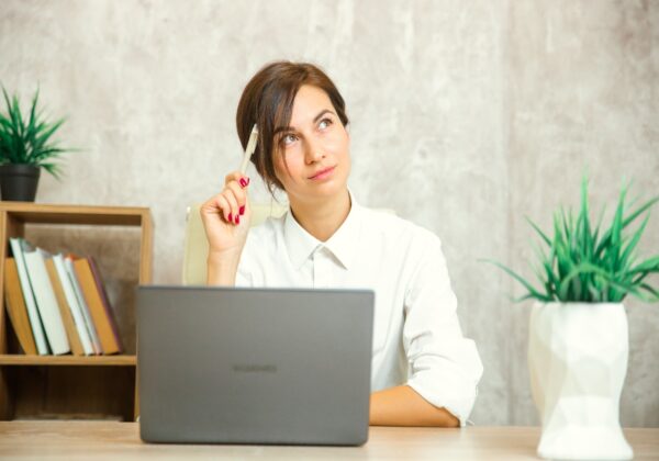Woman in white button-up shirt sitting at wooden desk with laptop, holding pen thoughtfully while looking up, surrounded by books and plants in modern office setting.