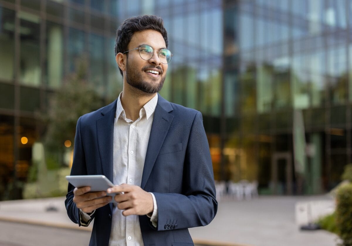 A confident professional smiles at the camera, arms crossed, outside a glass office building.