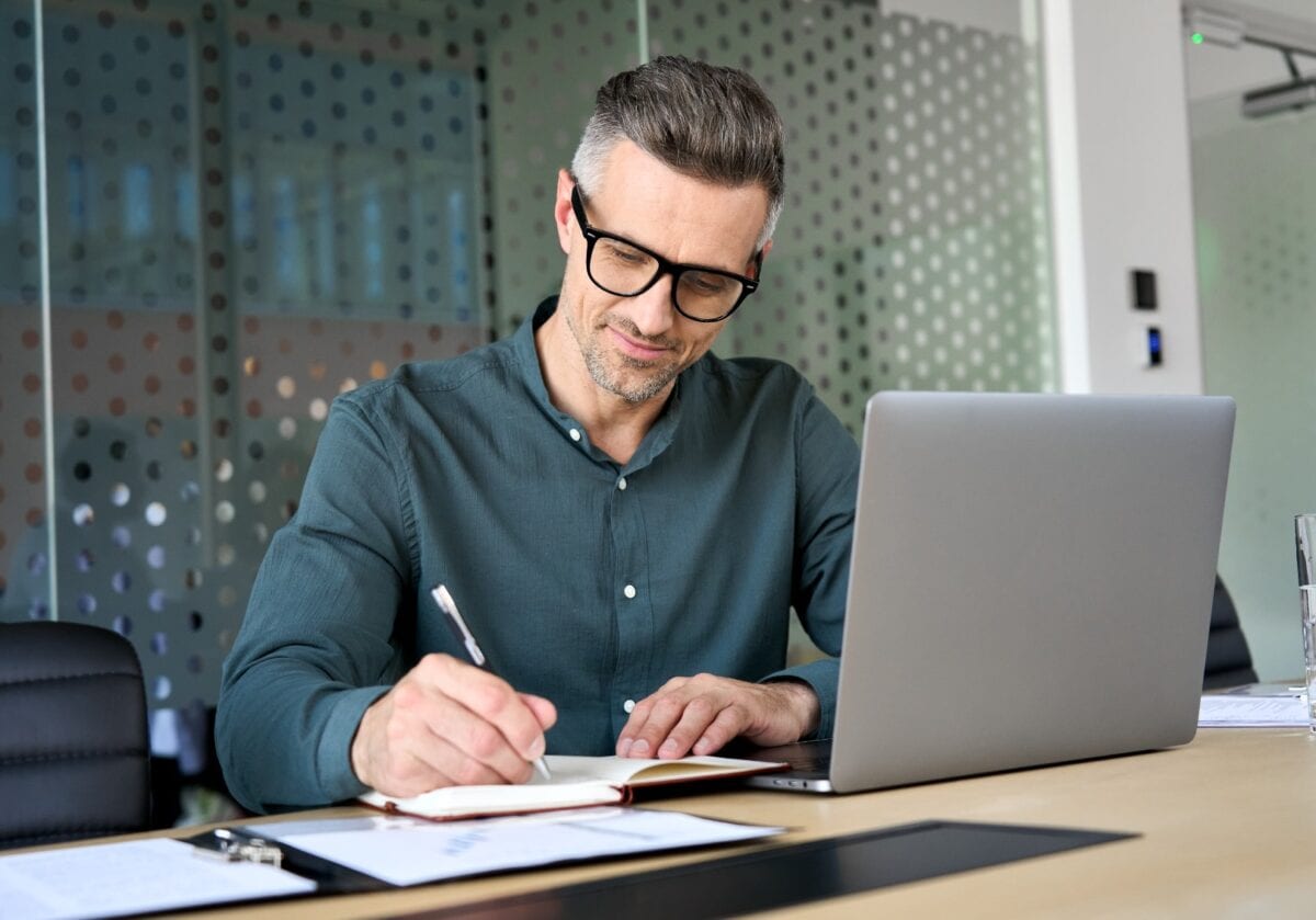 A focused, middle-aged businessman in glasses working at his desk, writing in a notebook next to a laptop.