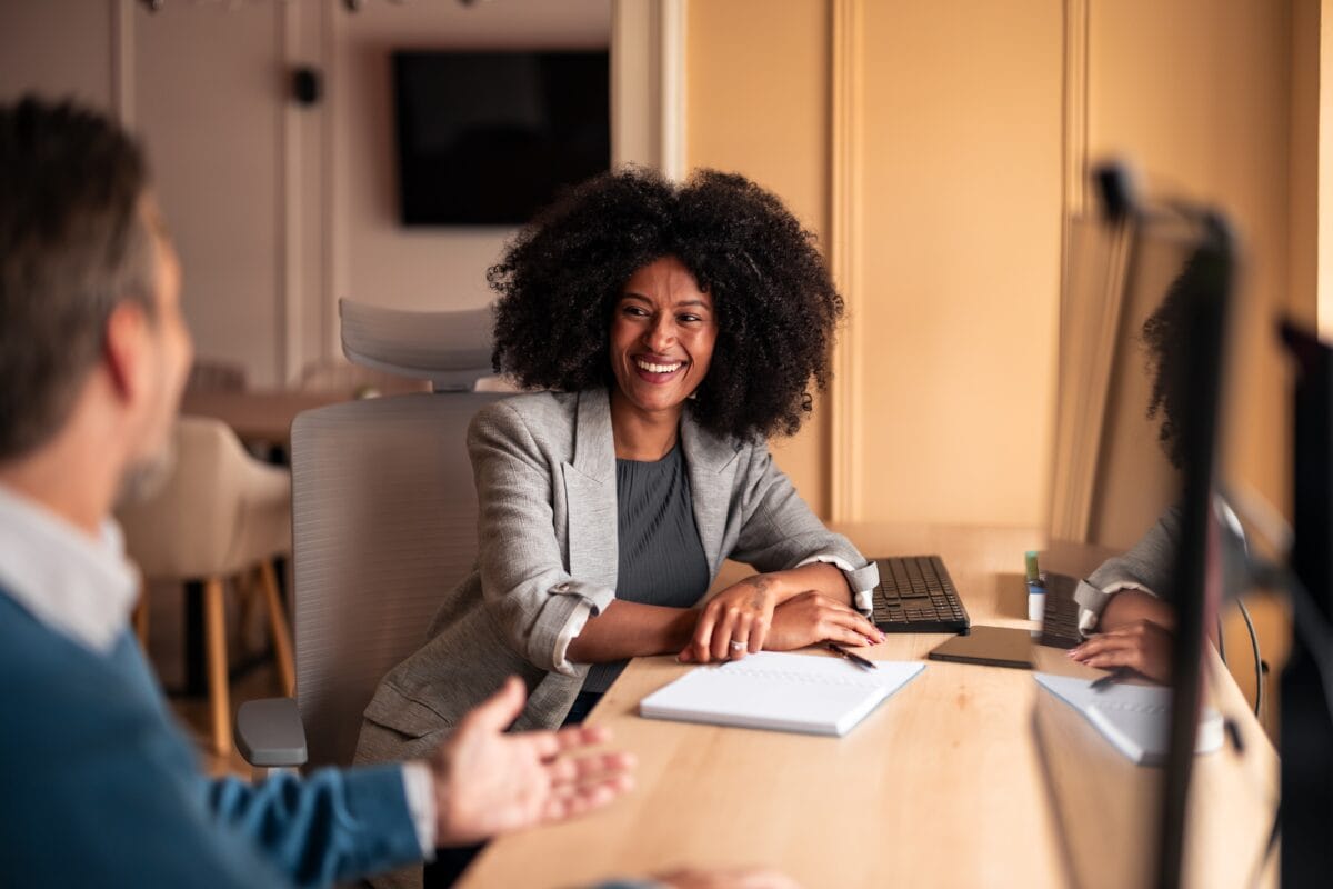 Smiling woman seated at a desk talking with a colleague in an office