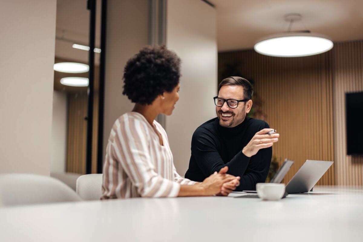 Two professionals smiling and talking across a table in a modern office setting