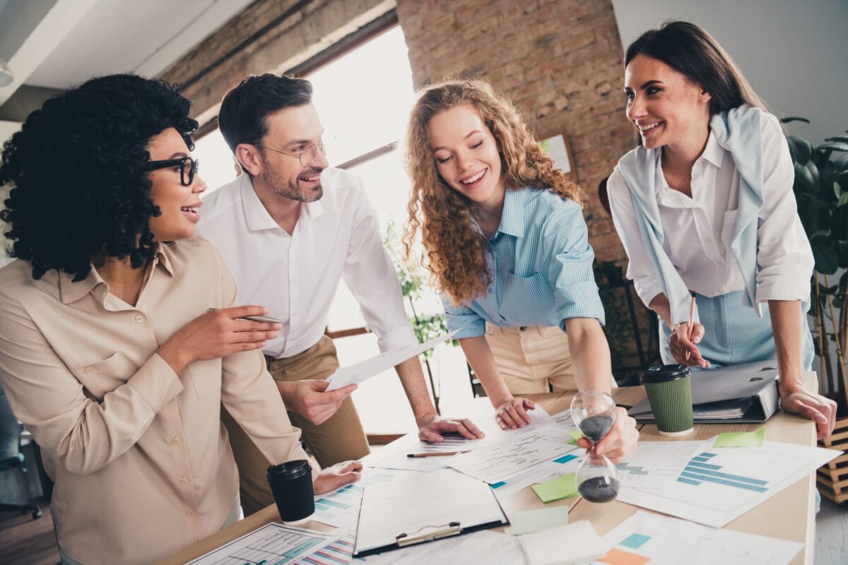 Four colleagues standing around table discussing charts and ideas in office