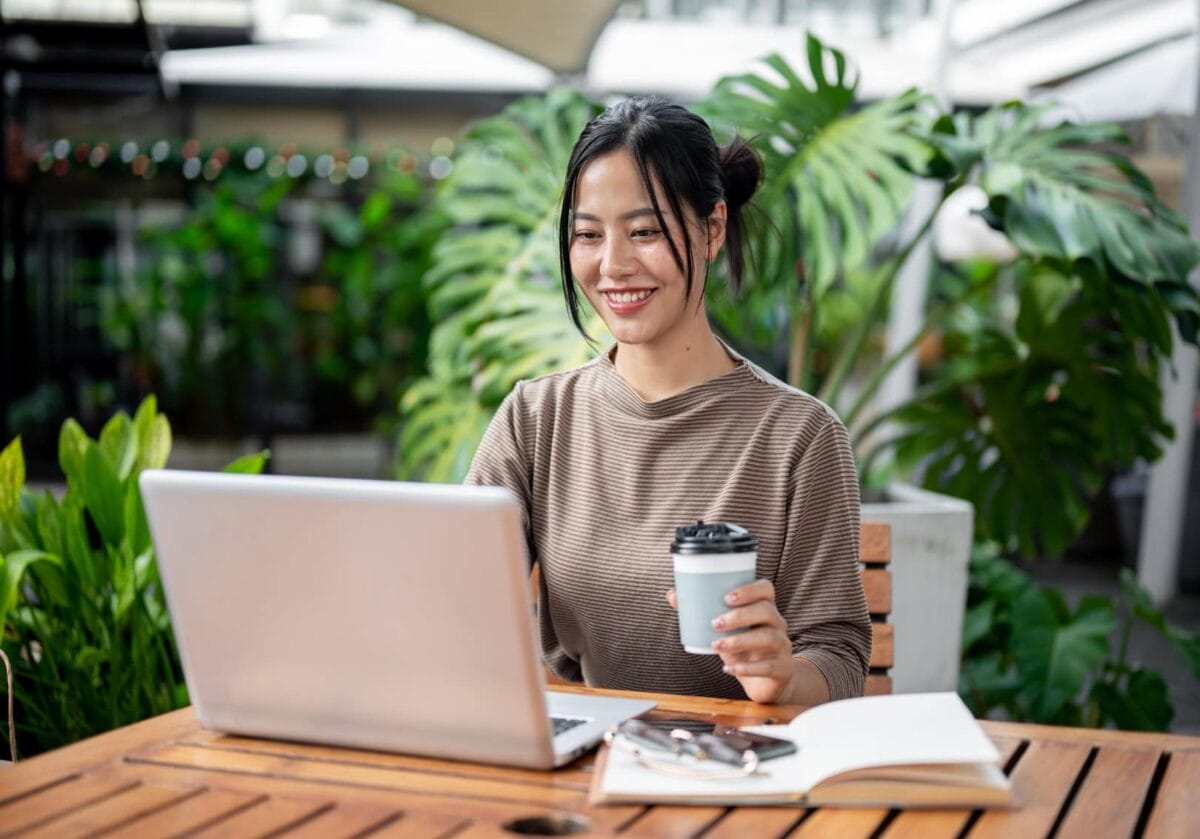 A smiling young woman sitting outdoors with a laptop, holding a coffee cup, surrounded by lush green plants.
