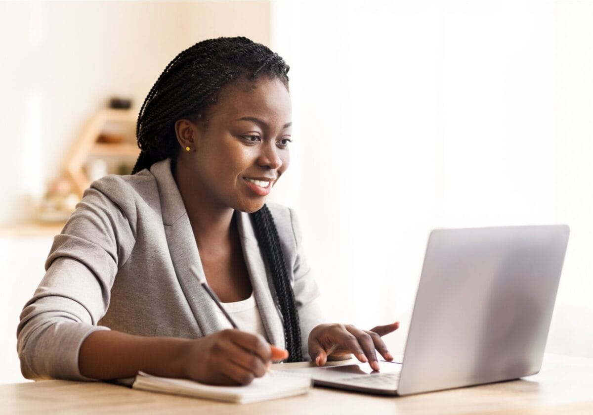 Smiling black professional writing in a notebook while looking at a laptop computer.