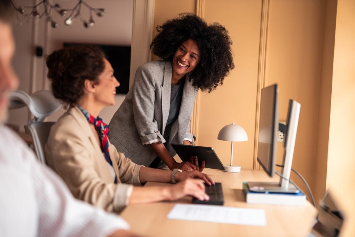 Two women are talking about becoming a coach while looking at resources on a computer, within a professional setting.