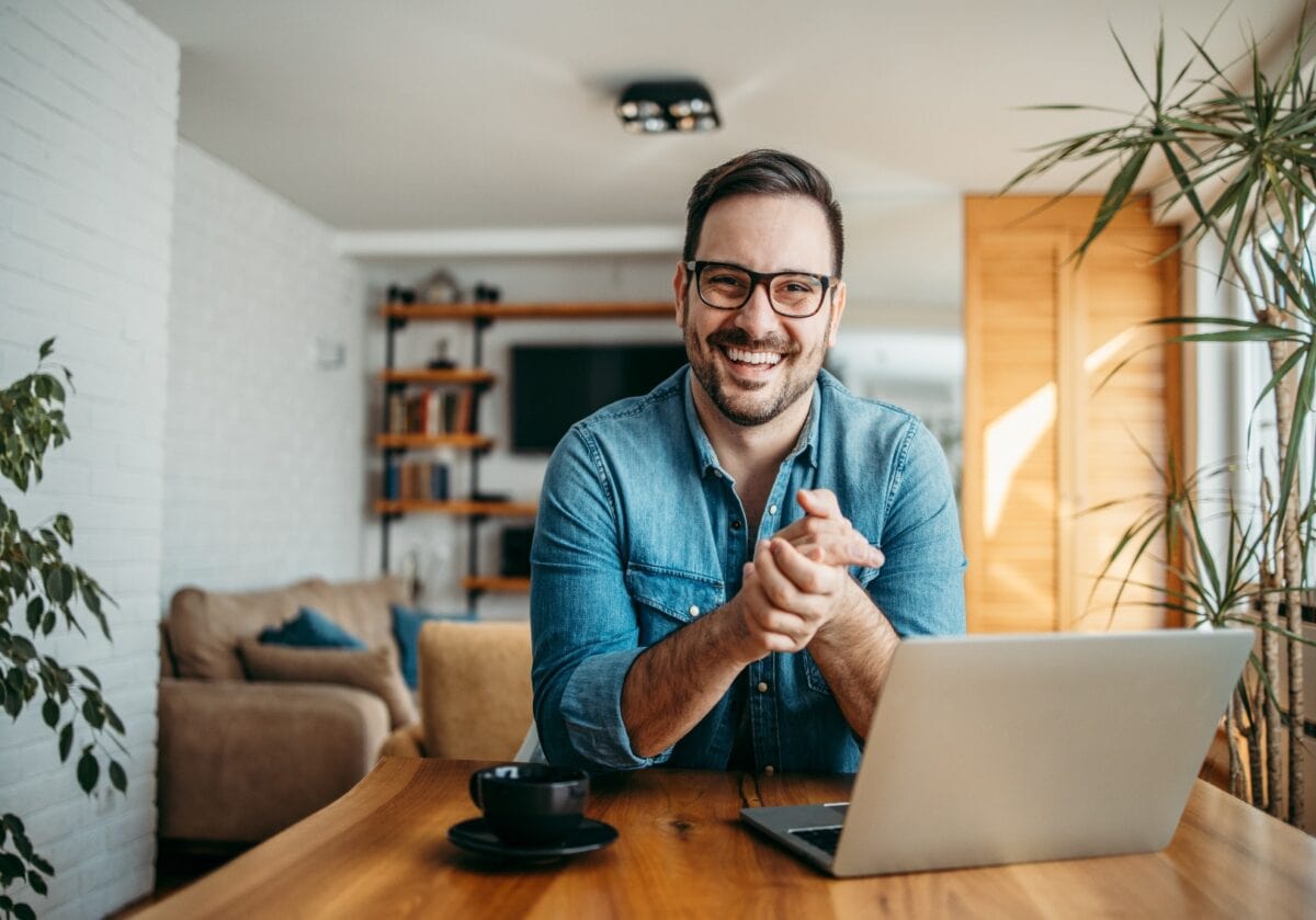 A smiling male professional sitting at a wooden table with a laptop, representing a modern, approachable coach working in a home office setting.
