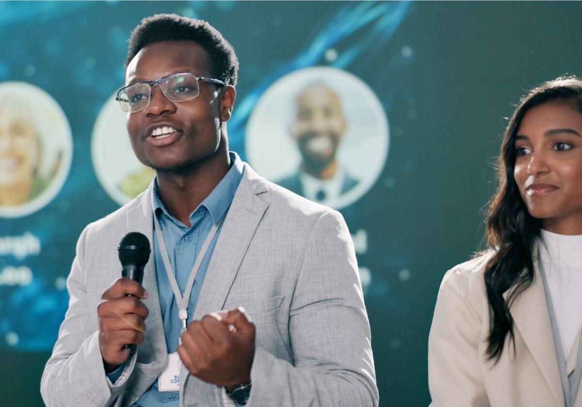 A man in a gray blazer speaks into a microphone at a professional conference while a woman beside him listens attentively.