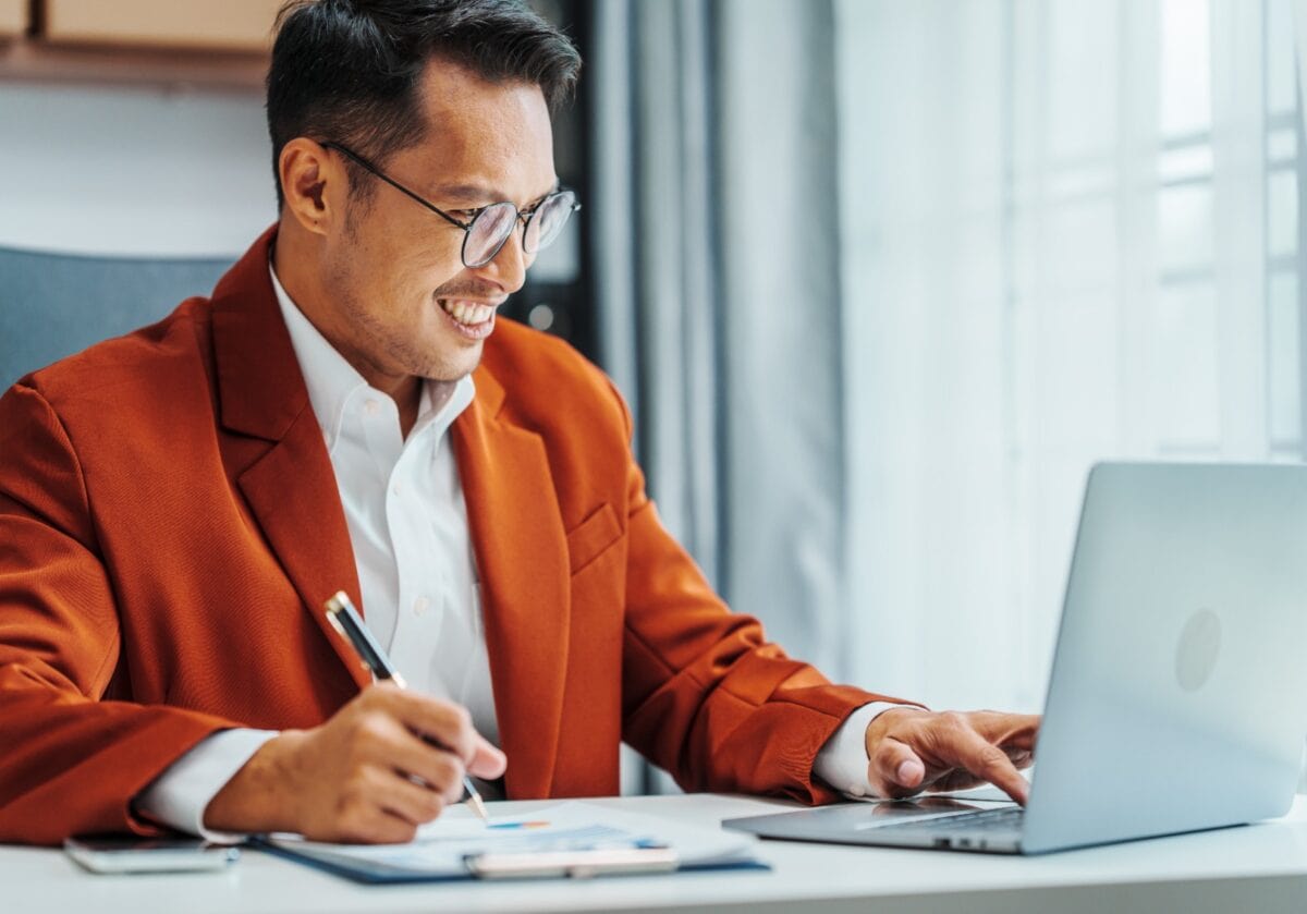 A smiling professional Asian Indian male in an orange blazer working on a laptop and taking notes.