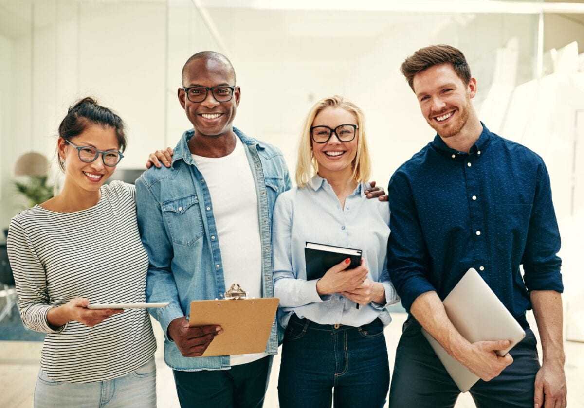 A diverse group of four smiling professionals standing together in a bright office, illustrating why hiring an executive coach can elevate your team and career.