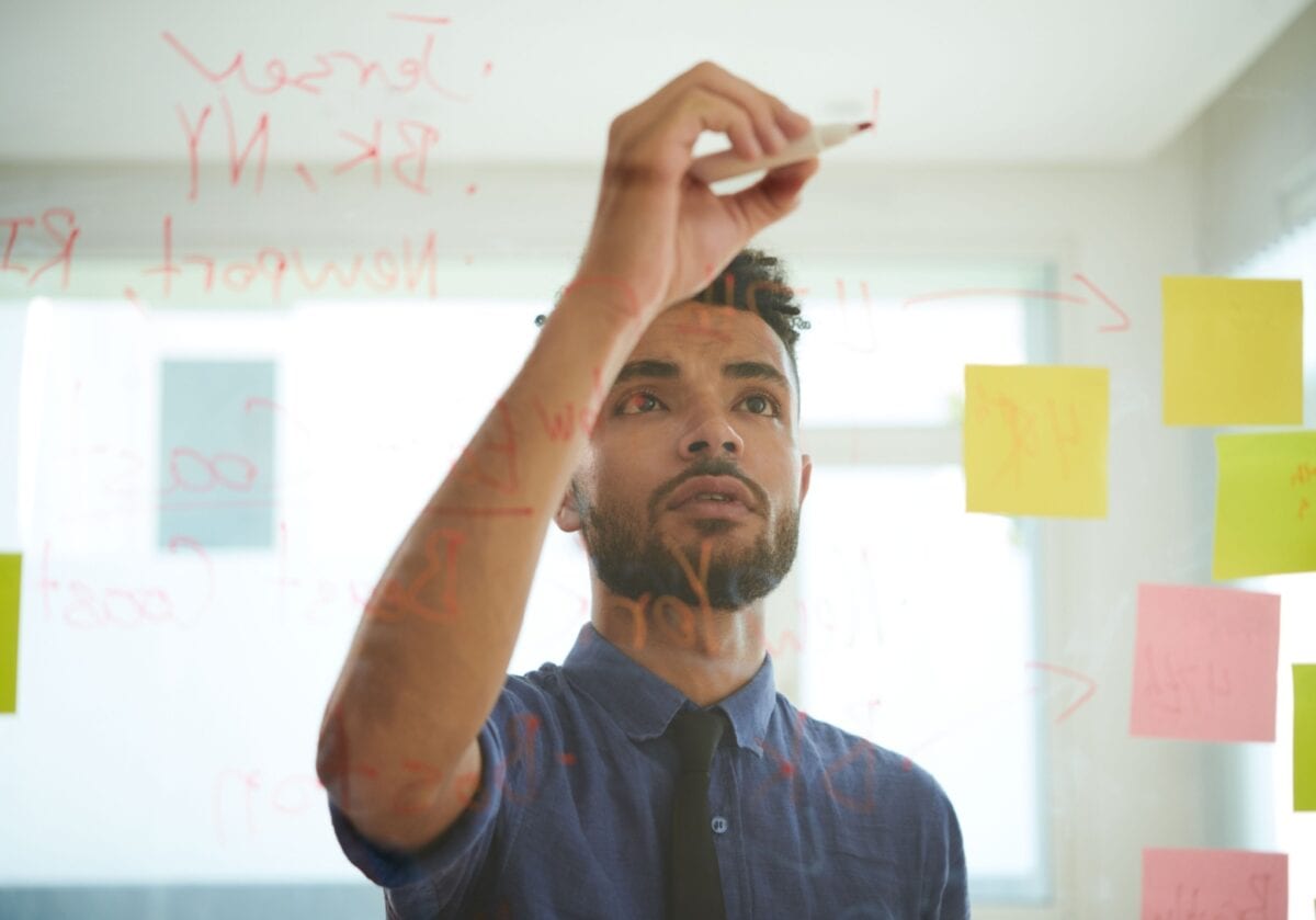 A professional man in a blue shirt and tie writing strategy notes and mapping ideas on a glass board with sticky notes.