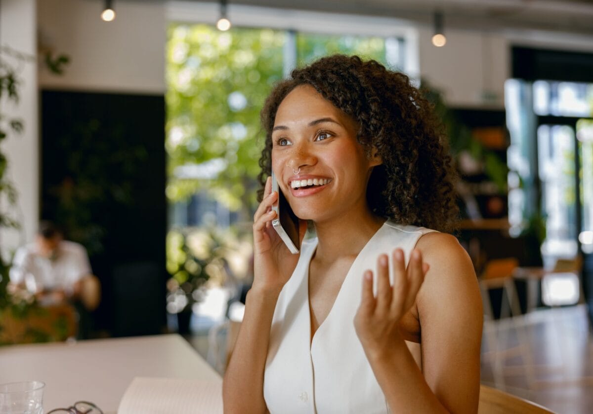 A smiling Black woman with curly hair talking on a smartphone in a bright, modern office or café setting.