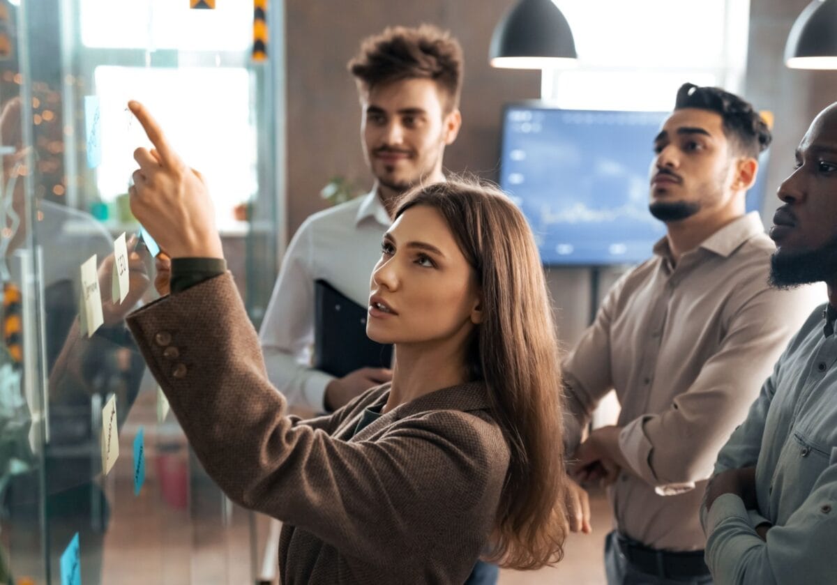 A group of professionals discusses ideas while one person points to notes on a glass wall.