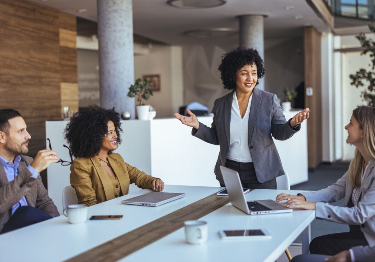 A business professional stands and speaks to three colleagues during a meeting in a modern office, while the team listens and smiles around a conference table with laptops.